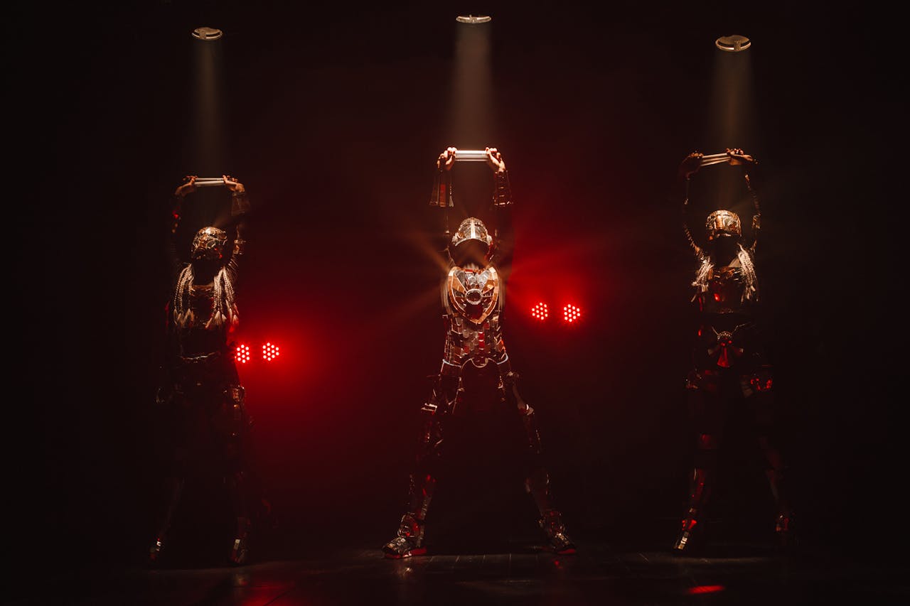 Three performers in elaborate costumes on stage, illuminated by dramatic red lighting.